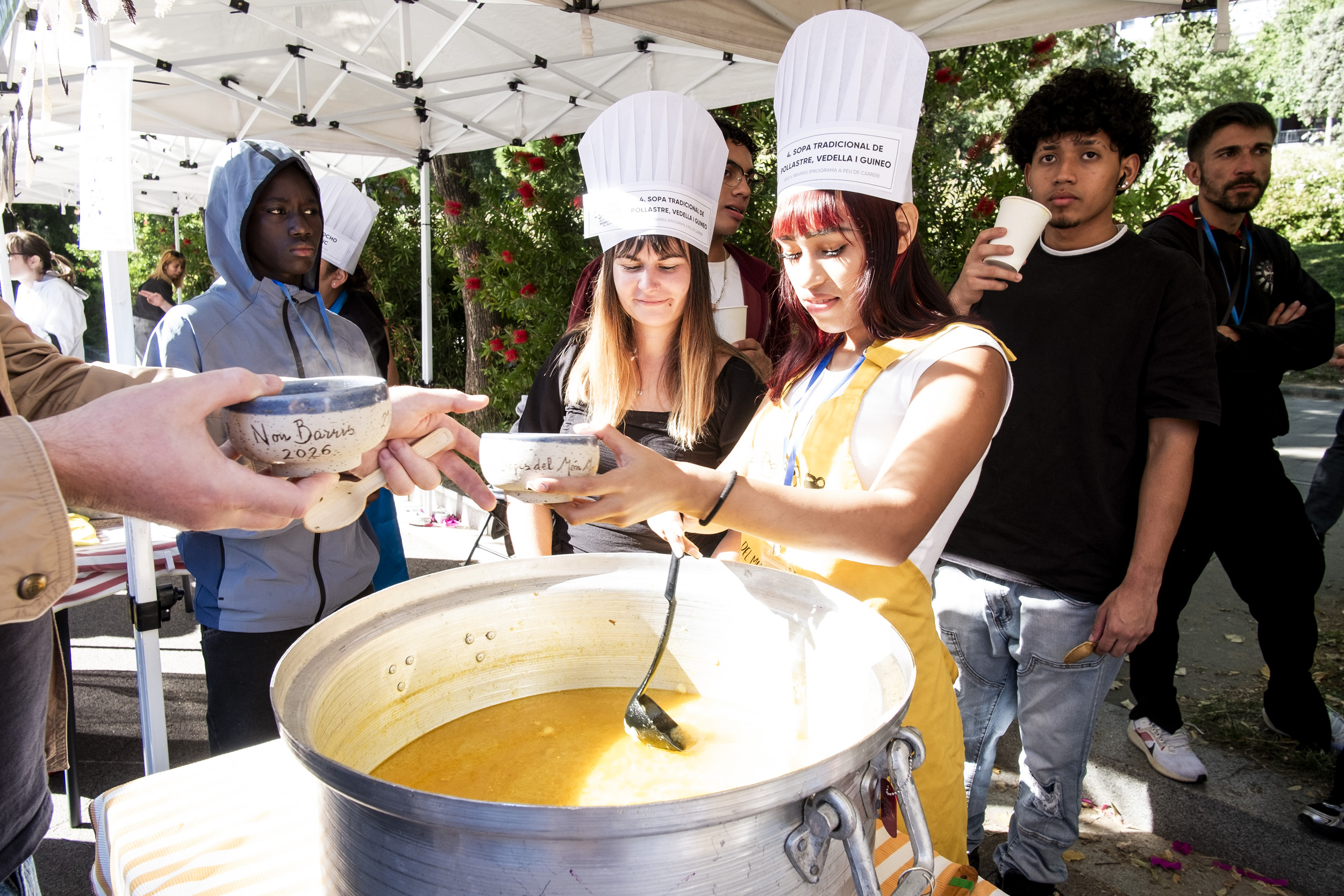 Joves participants reparteixen la seva sopa durant el 21è Festival de Sopes del Món Mundial de Nou Barris, celebrat al barri de Canyelles el 25 d’octubre de 2026, en una jornada de convivència i diversitat cultural.